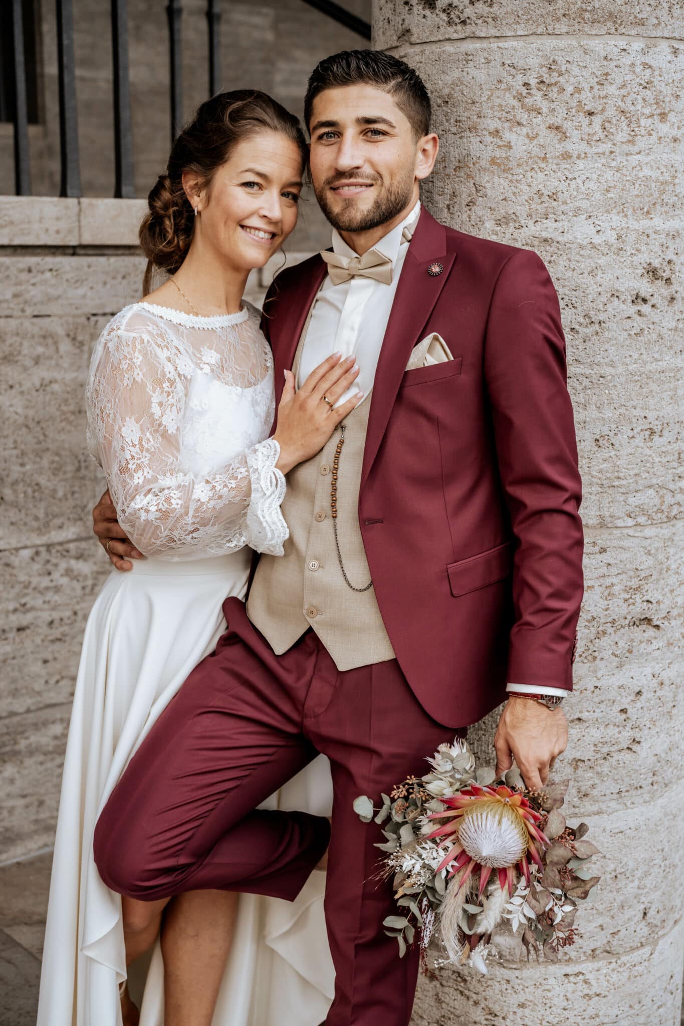 Braut und Bräutigam lächeln und posieren in formeller Hochzeitskleidung, der Bräutigam hält einen Brautstrauß.